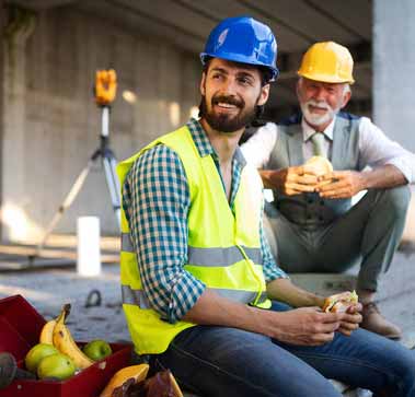 Zwei Bauarbeiter sitzen auf einer Baustelle und essen Brötchen. Sie machen Pause.