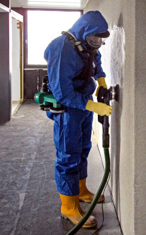 Ein Mann in Schutzanzug und Atemschutzmaske entfernt den Putz von der Wand mit einem Bearbeitungssystem, das mit Absaugung ausgestattet ist.