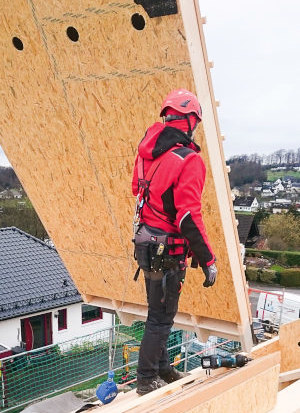 Person mit Helm, Schutzkleidung und persönlicher Schutzausrüstung gegen Absturz schiebt auf einem Dach ein hängendes Holzbauelement weg.