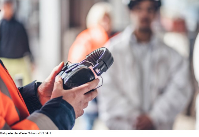 Eine Person in Warnkleidung hält einen Gehörschutz in der Hand.