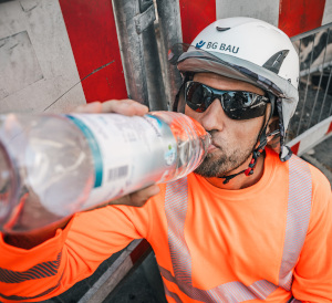 Ein Bauarbeiter in Warnkleidung und Schutzhelm trinkt an einer Baustelle bei heißem Wetter Wasser, um sich abzukühlen.