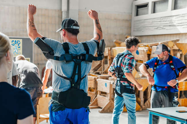 Ein Auszubildender trägt ein Exoskelett und hebt die Arme. Im Hintergrund stehen weitere Menschen mit weiteren Exoskeletten.