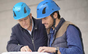 Zwei Bauarbeiter mit Schutzhelmen schauen auf einen Bauplan und beraten sich.