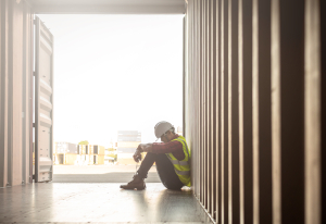Ein Arbeiter sitzt mit angewinkelten Beinen in einem offenen Container. Der Kopf ist nach unten gebeugt.