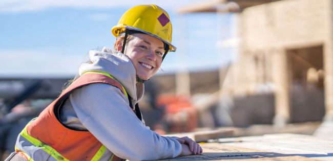 Eine Bauarbeiterin in Arbeitskleidung lehnt auf einer Palette mit Holzbohlen und lächelt in die Kamera. Im Hintergrund ist eine Baustelle erkennbar. 