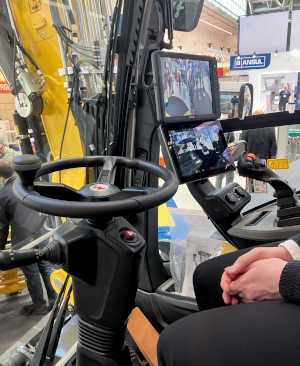 Innenaufnahme einer Baggerkabine, die in einer Messehalle aufgestellt ist. Neben dem Lenkrad sind zwei Monitore zu sehen, die das Umfeld über eine Kamera zeigen.