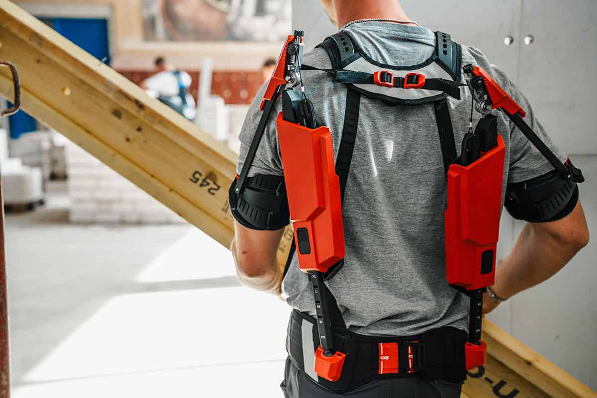 Ein Auszubildender trägt mit einem Exoskelett einen Holzbalken.