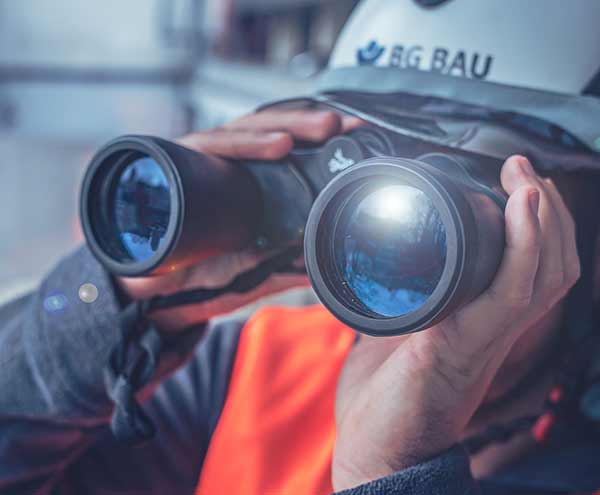 Eine Person mit BG BAU-Schutzhelm blickt durch ein Fernglas.