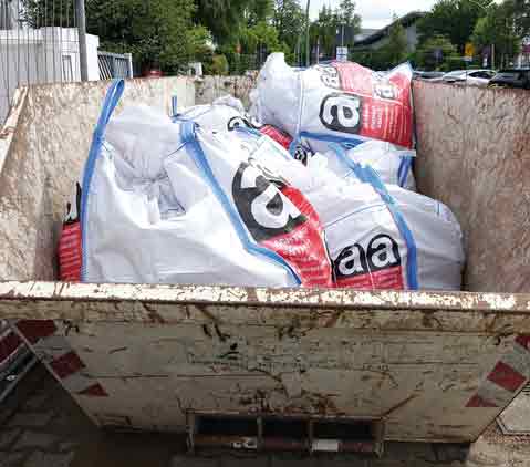 Ein Metallcontainer auf einer Baustelle am Straßenrand mit weißen Säcken, die mit Warnhinweisen für asbesthaltige Abfälle gekennzeichnet sind.