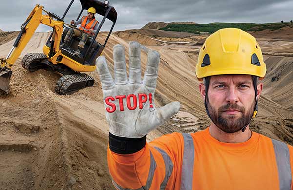 Ein Bauarbeiter hebt seine Hand, auf deren Handschuh "Stop" steht, während eine Bauarbeiterin in einem Bagger in Schieflage auf Sand gerät.