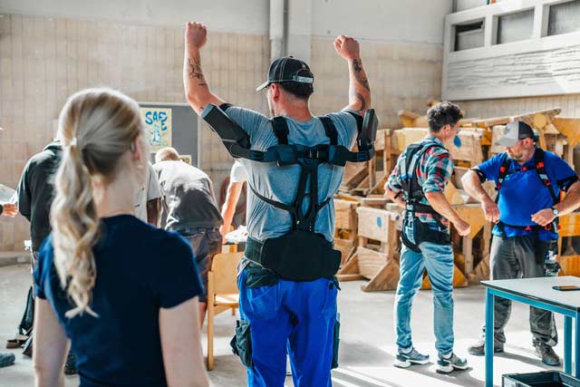 Ein Auszubildender trägt ein Exoskelett und hebt die Arme. Im Hintergrund stehen weitere Menschen mit Exoskeletten.