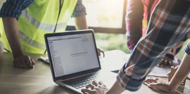 Drei Bauarbeiter stehen um einen Laptop herum und schauen auf den Laptop-Bildschirm.